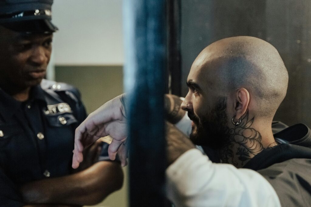 Close-up of a police officer interacting with a tattooed man behind metal bars inside a jail.