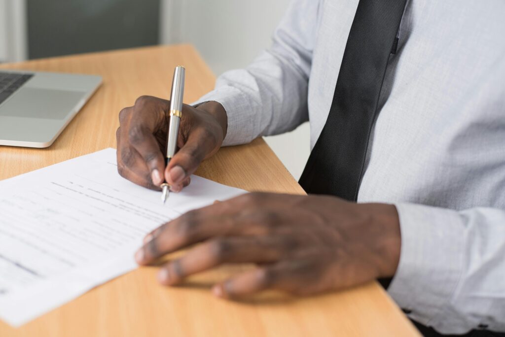 Close-up of a man signing paperwork on a desk, with a laptop nearby, in a professional office environment.